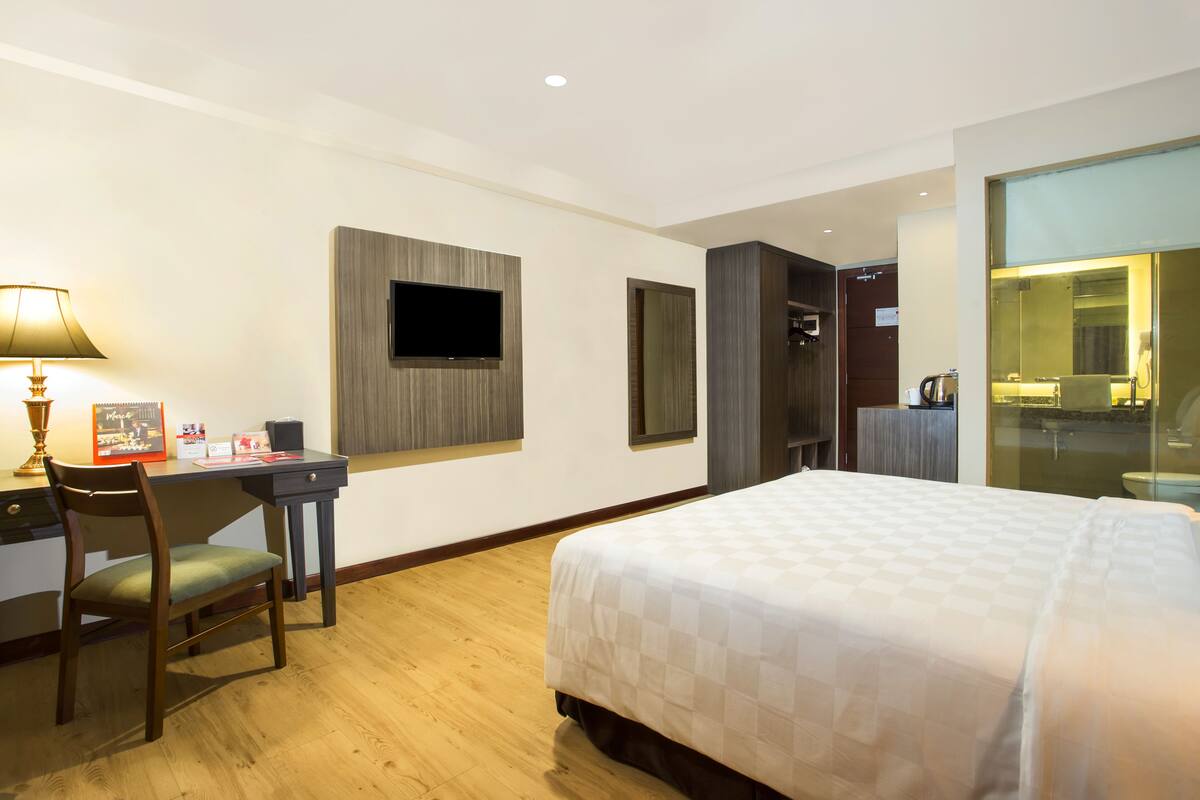A hotel room with a bed, desk, chair, TV, and a bathroom visible through a glass door.