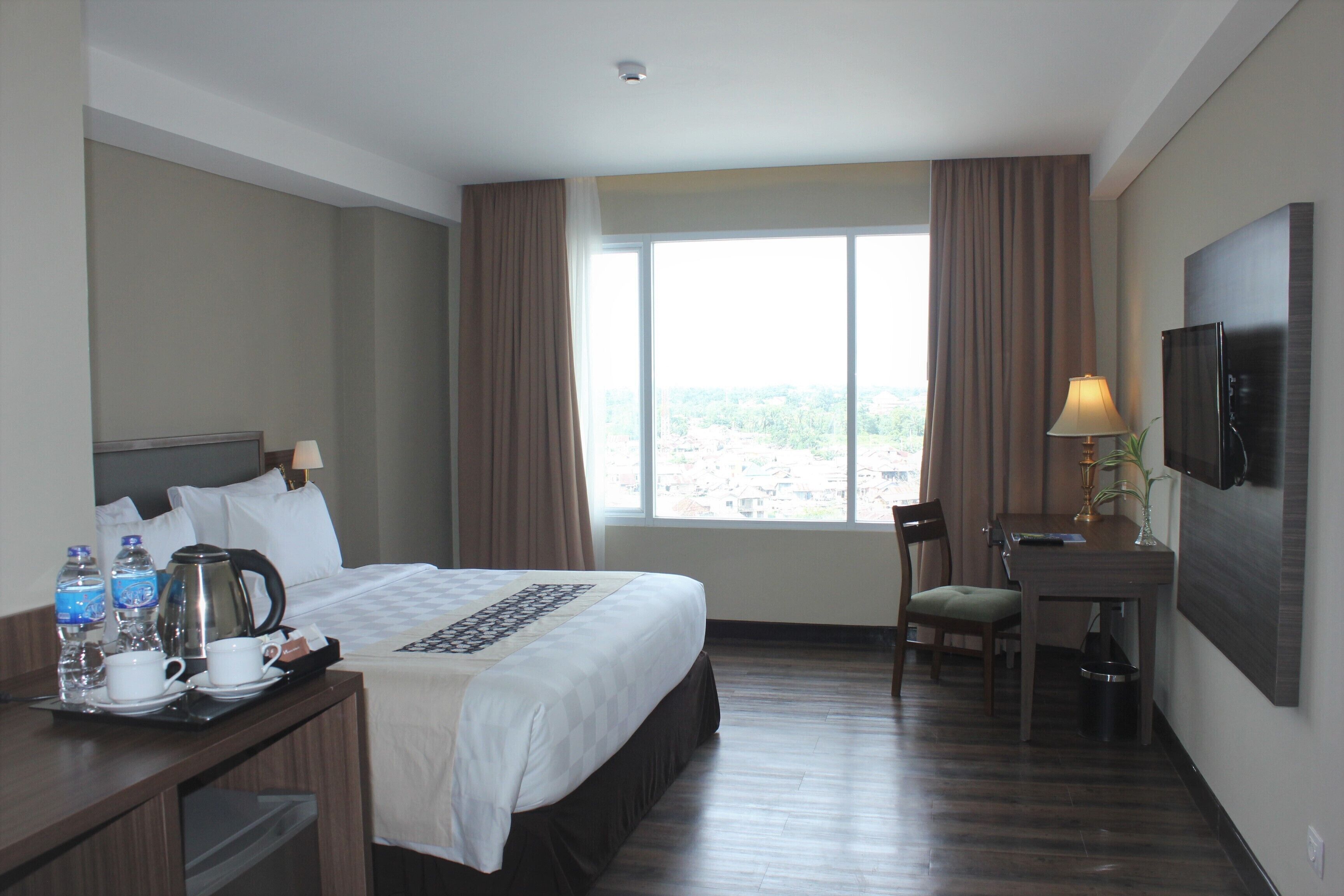 Une chambre d’hôtel avec un lit, un bureau avec une lampe, une télévision et une fenêtre avec des rideaux.