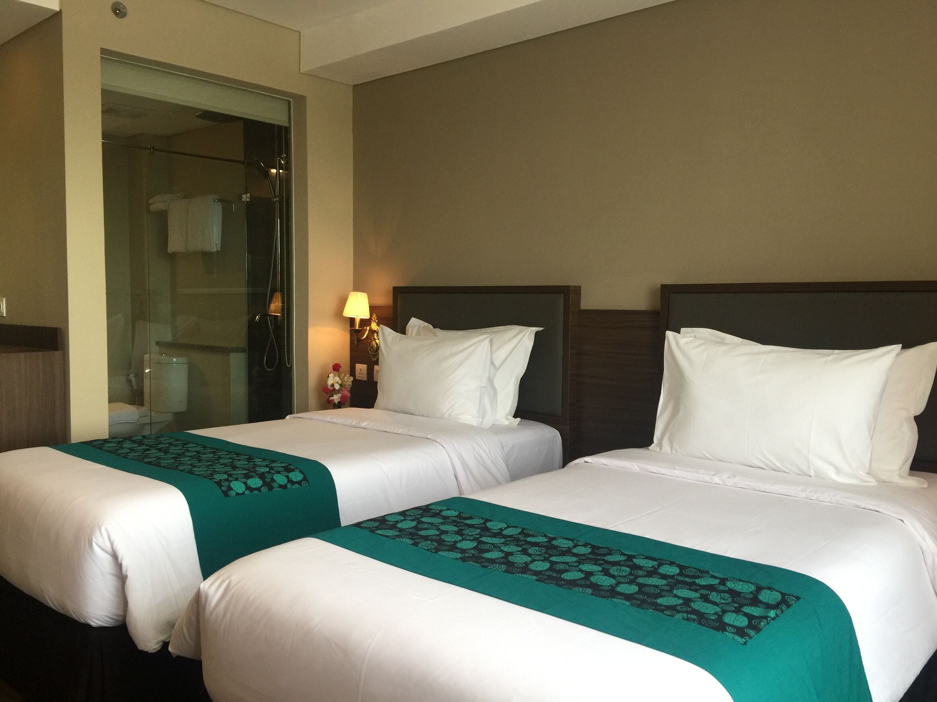Ein Hotelzimmer mit zwei Betten, einer Glasdusche, einem Nachttisch mit Lampe und einer Blumenkomposition.