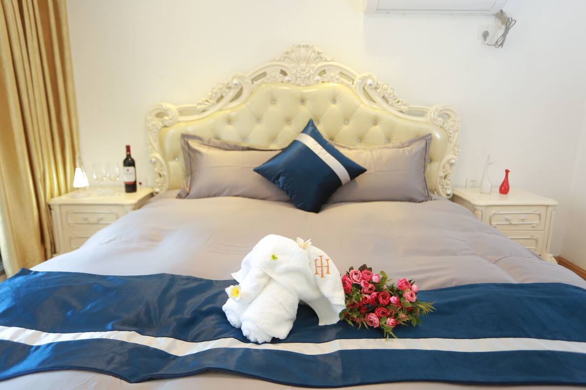 Un lit avec une tête de lit capitonnée, une couvre-lit bleue et blanche, un éléphant en peluche blanc et un bouquet de fleurs roses.
