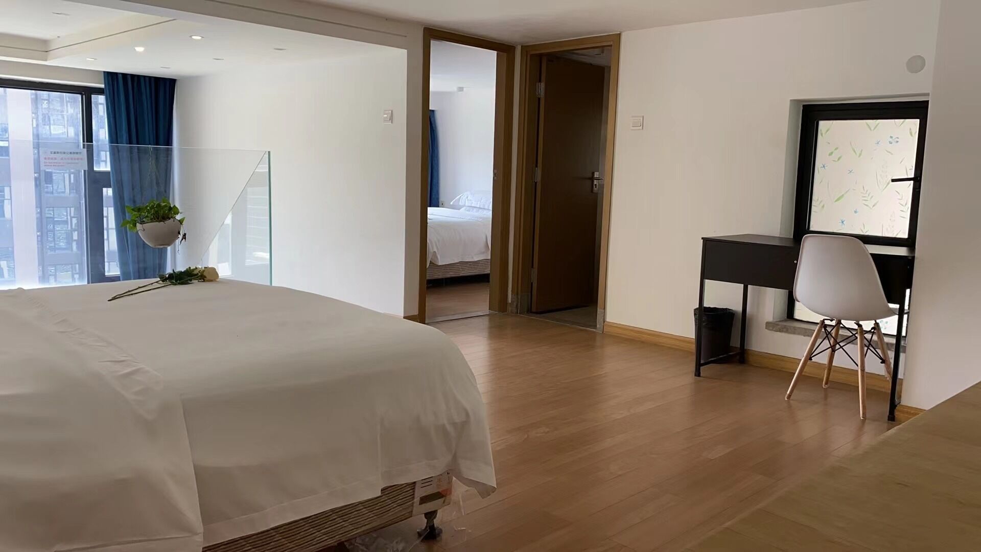 Ein modernes Hotelzimmer mit einem großen Bett, einem Schreibtisch und einem Stuhl. Ein Fenster bietet Blick auf Gebäude, und eine Tür führt in einen weiteren Raum.
