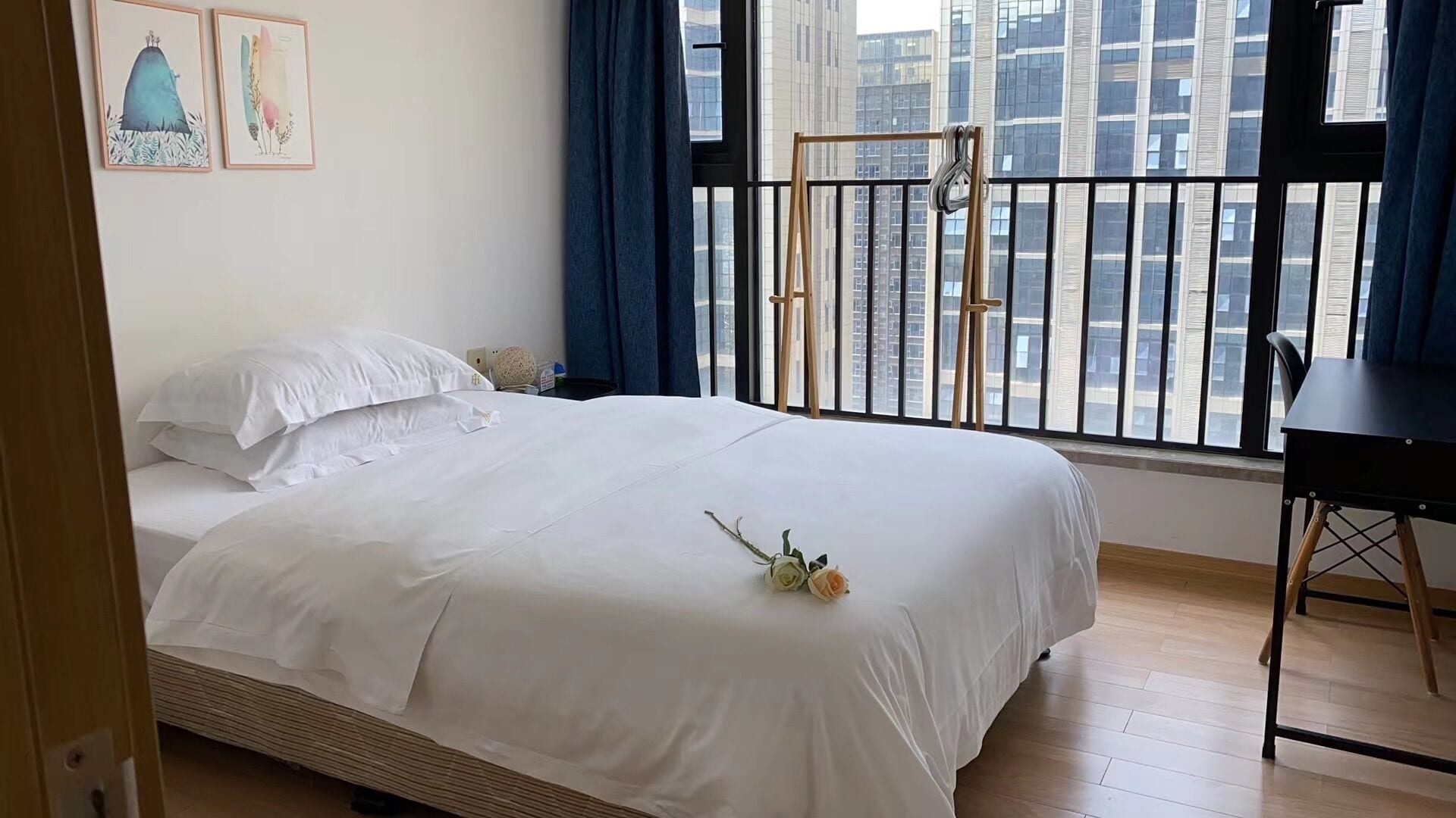Un lit bien fait, avec du linge de lit blanc, une petite table de chevet avec une lampe, et une vue sur le paysage urbain depuis un balcon doté d’une rambarde noire.