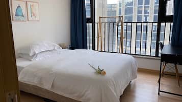 A neatly made bed with white linens, a small bedside table with a lamp, and a view of a cityscape through a balcony with a black railing.