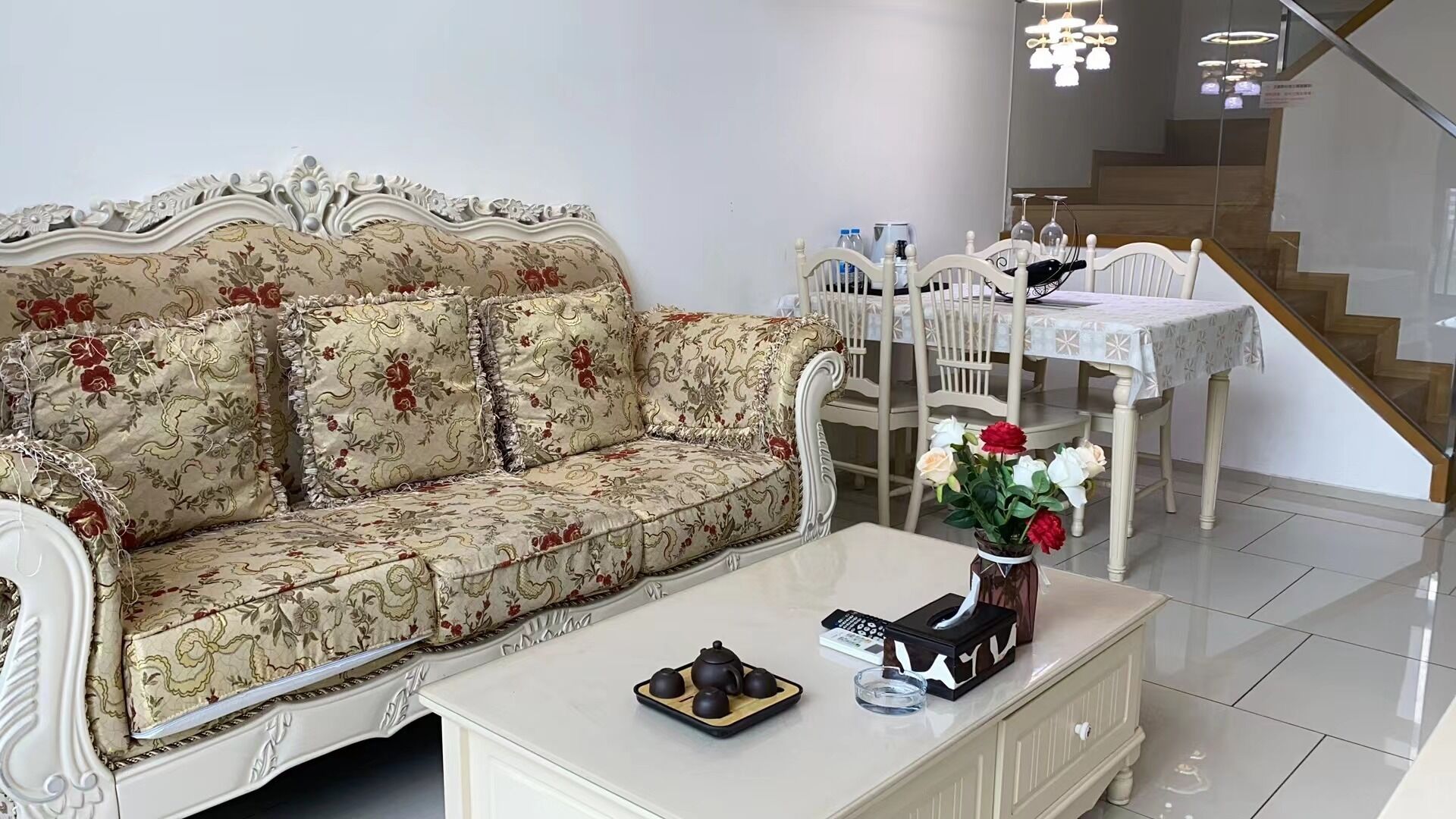 En stue med en blomstret sofa, et hvidt sofabord med tepotte og kopper, et spisebord med stole, og en trappe i baggrunden.