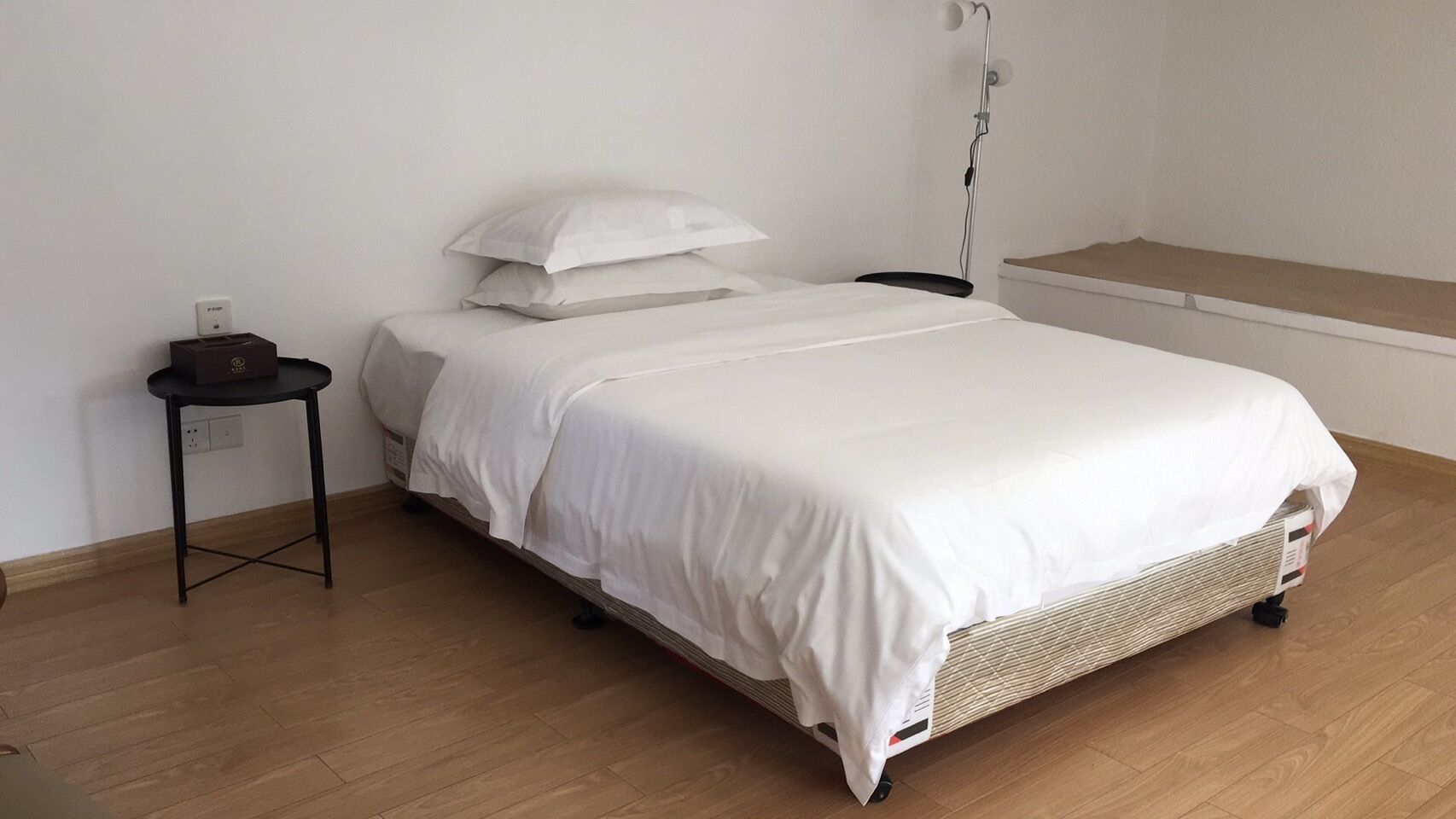 A neatly made bed with white bedding, a small bedside table, and a wall-mounted lamp.