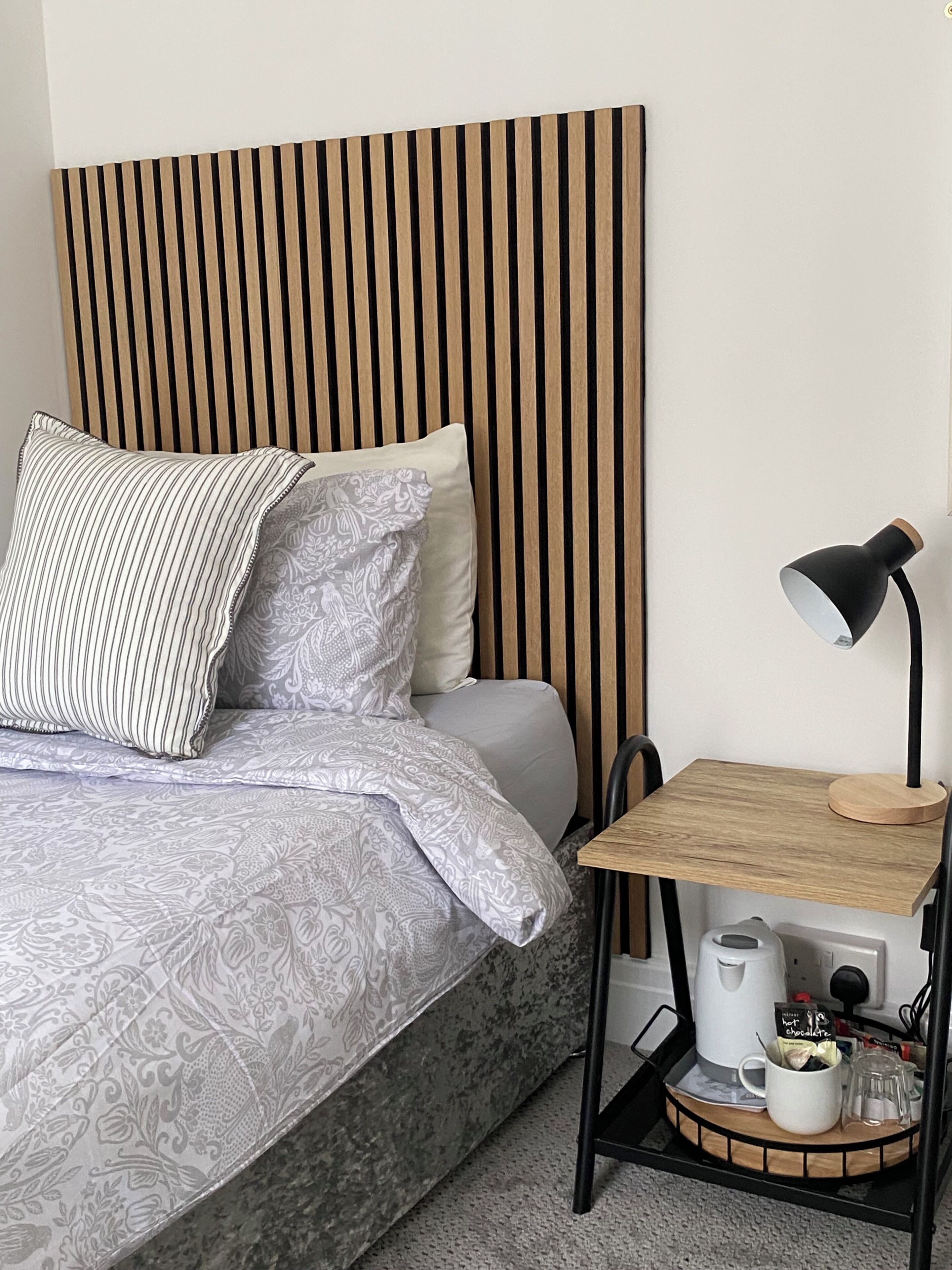 Habitación de hotel ordenada con cama, mesita de noche con lámpara, cafetera y bandeja con taza y taza de café.