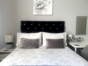 A bedroom with a tufted headboard, two pillows, a bedside table with a lamp, a mirror, and a tray with toiletries. - Brene Hotel (Blackpool)