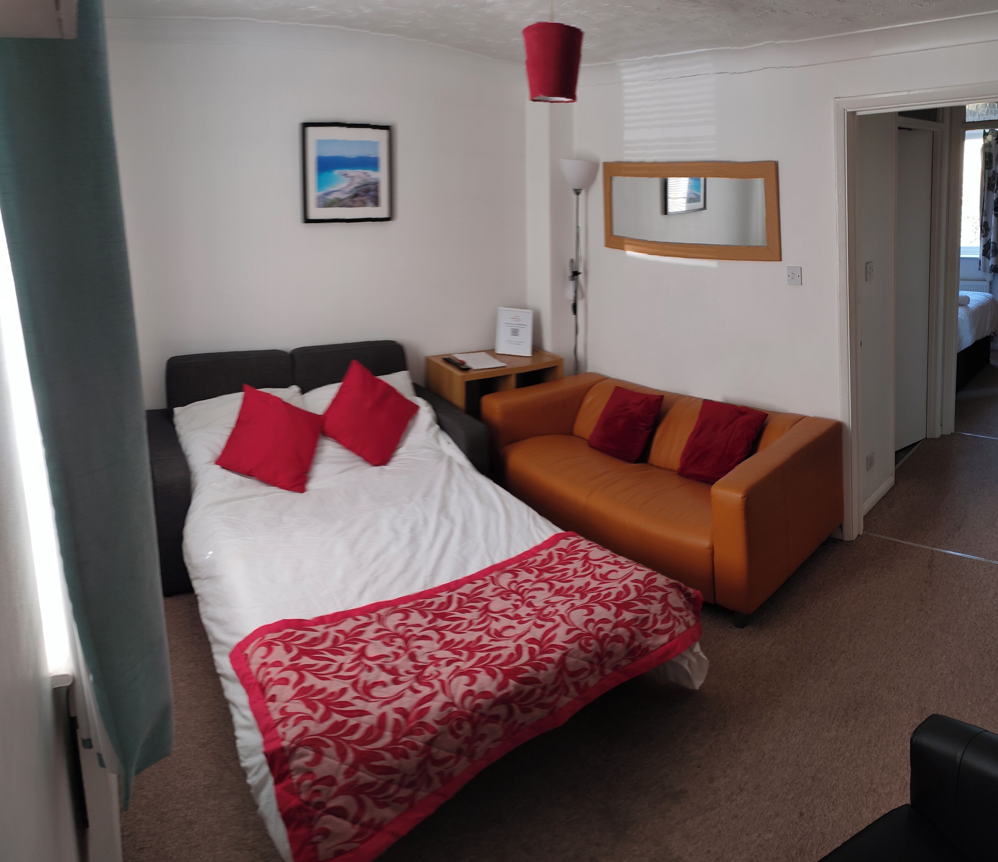 A hotel room with a bed, a sofa, a bedside table, a lamp, a mirror, and a framed picture on the wall.