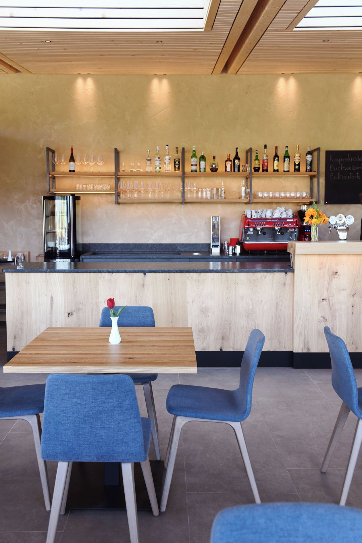A modern bar area with wooden furniture, a variety of bottles and glasses on shelves, and a chalkboard menu.