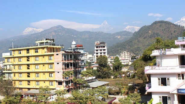 A multi-story building with a yellow facade, surrounded by other buildings and greenery, with a mountainous backdrop. - Hotel Lake Valley (Pokhara)