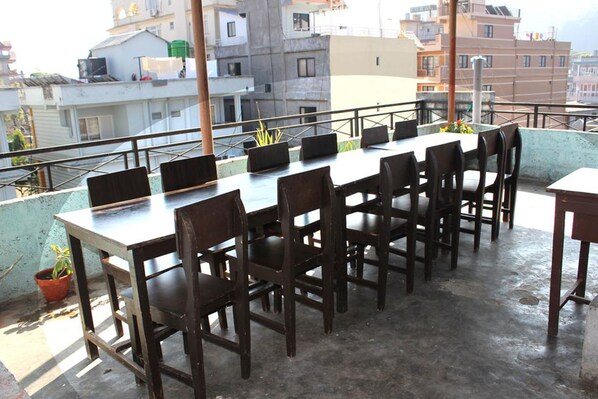 A rooftop terrace with a long table and chairs, surrounded by a metal railing, overlooking a residential area.