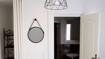 Une chambre d’hôtel avec deux lits, un banc, un miroir rond, un luminaire suspendu et une salle de bain visible à travers une porte ouverte.