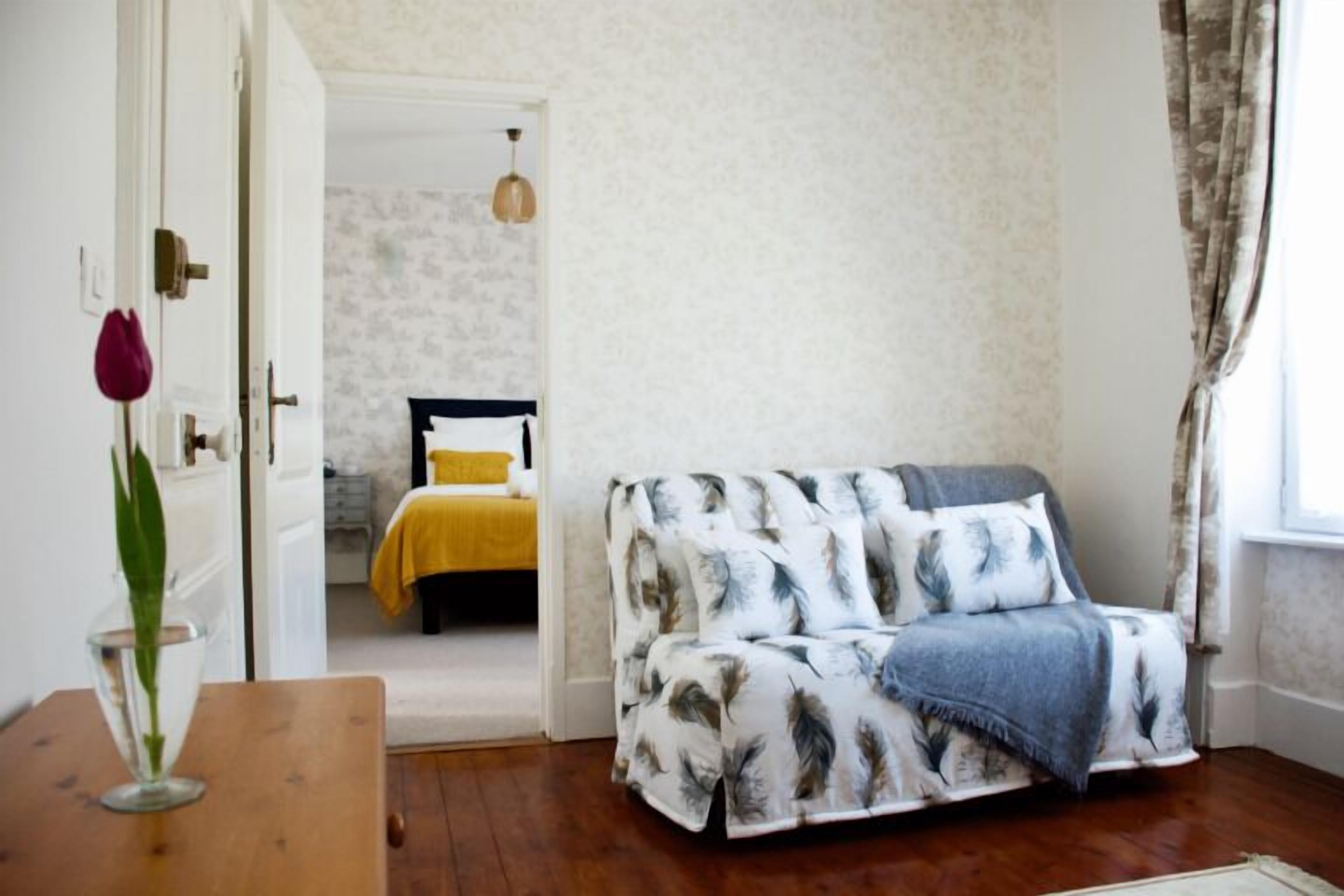 Una stanza con un divano rivestito di un motivo a foglie, un tavolo di legno con un vaso e un letto con una coperta gialla sullo sfondo.
