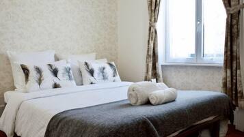 A neatly made bed with white and gray bedding, a pair of rolled towels, and a window with curtains.