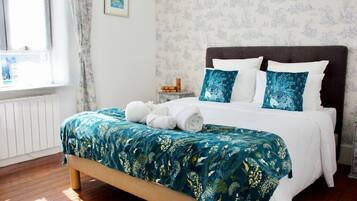 Cama bem arrumada com colcha florida, mesa de cabeceira com abajur e janela com cortinas.