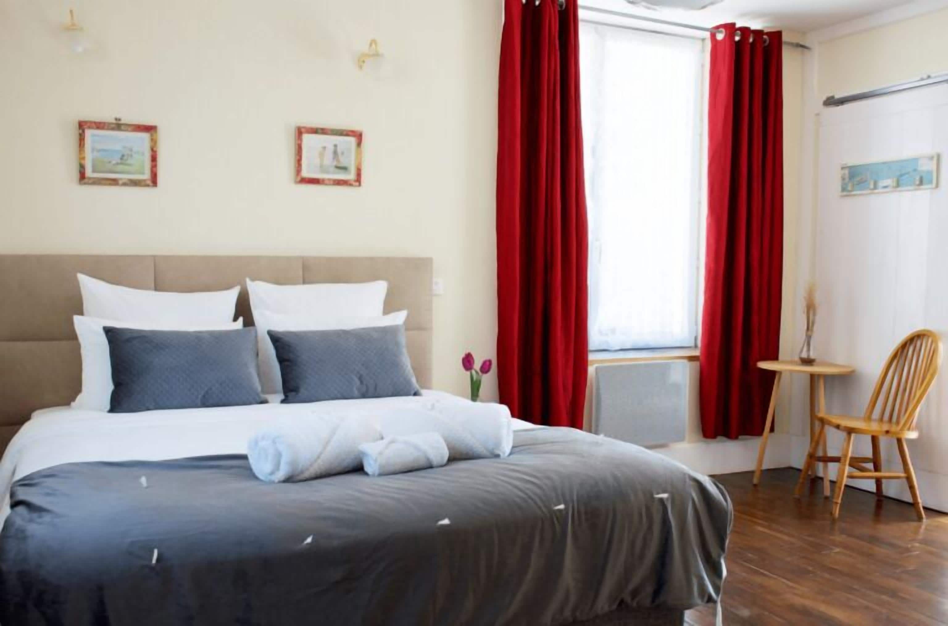 A neatly made bed with a grey and white duvet, red curtains, a small wooden table, and a chair.
