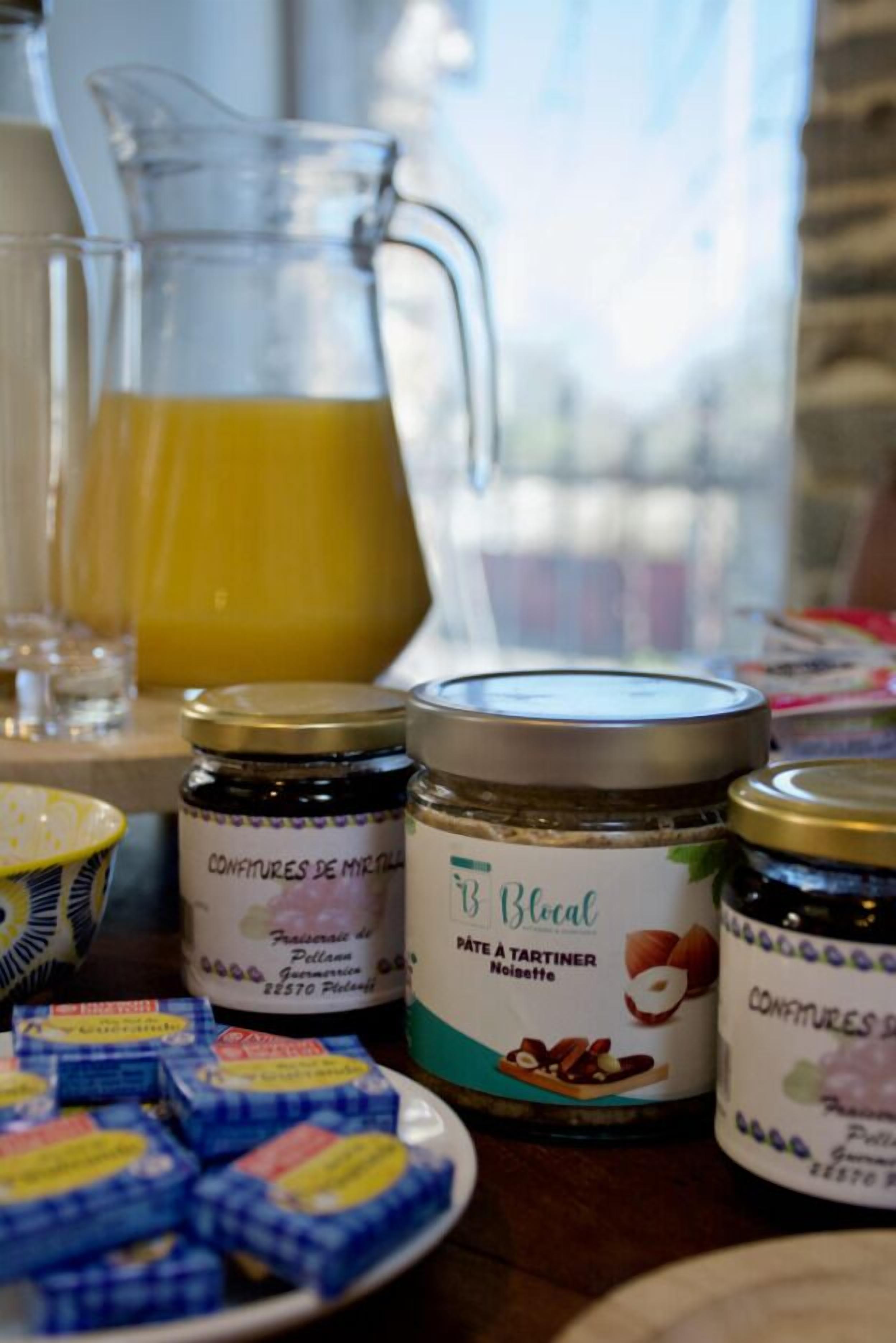 Una mesa con frascos de 'Confitures de Myrtilles' y 'Pâte à tartiner Noisette', una jarra de jugo de naranja y un plato con productos envasados en azul y amarillo.