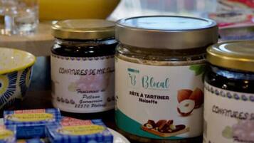 Una mesa con frascos de 'Confitures de Myrtilles' y 'Pâte à tartiner Noisette', una jarra de jugo de naranja y un plato con productos envasados en azul y amarillo.