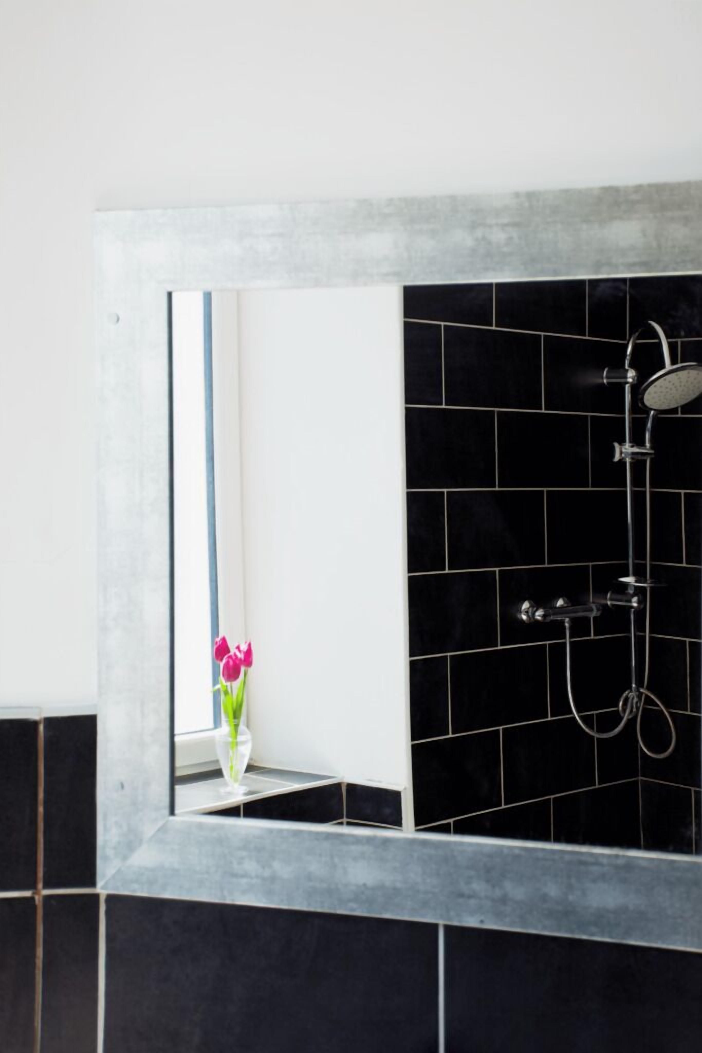 Een badkamer met zwart betegelde wanden, een witte wastafel en een spiegel die een raam met een bloevas weerkaatst.