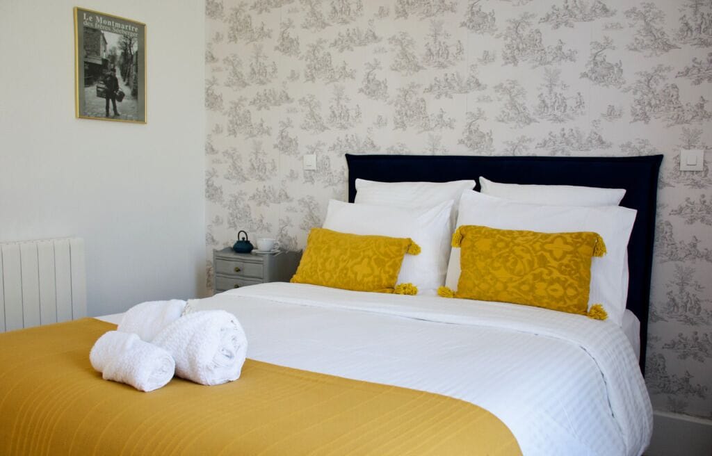 A neatly made bed with white and yellow bedding, a nightstand with a lamp, and a framed picture on the wall.