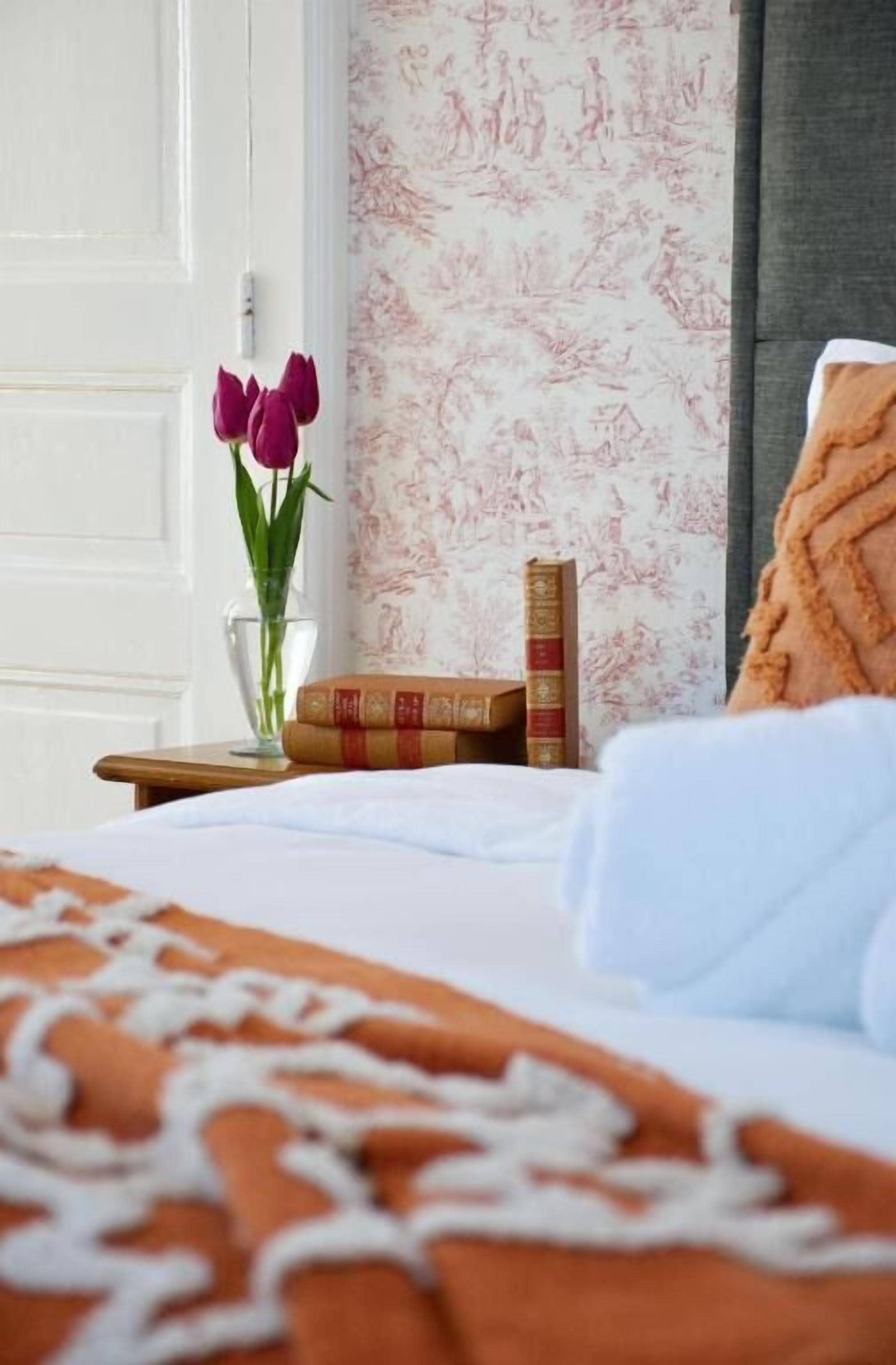 Ein ordentlich bezogenes Bett mit weiß-orangener Decke, eine Vase mit rosa Blumen und zwei Bücher auf einem Nachttisch.