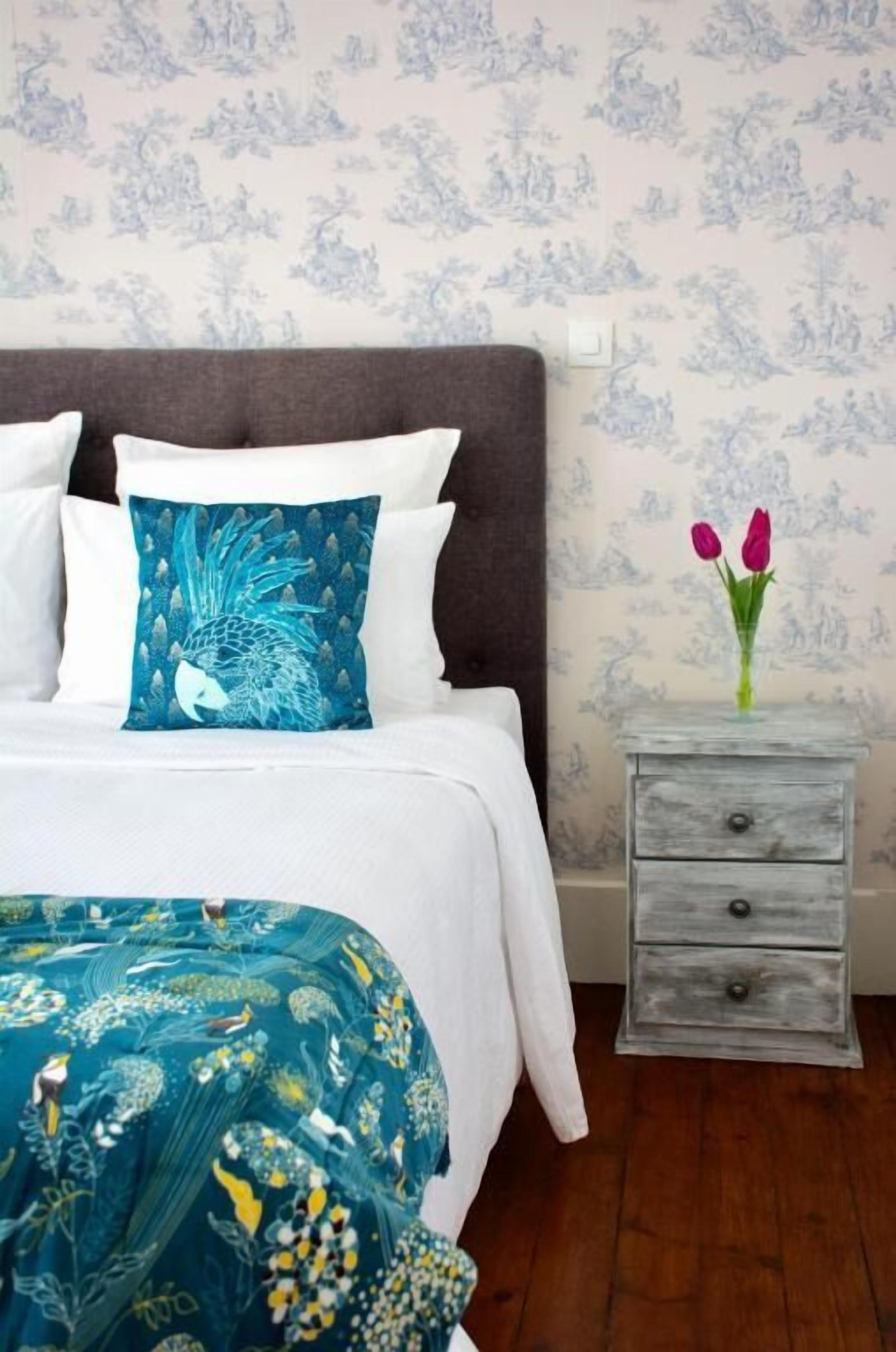 A neatly made bed with white bedding and a blue and white patterned pillow. A small wooden nightstand with two drawers and a vase of flowers is beside the bed.