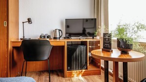 Kompakti hotellihuone, jossa on puinen työpöytä, musta tuoli, televisio, pieni jääkaappi, kahvinkeitin, ruukkukasvi ja pieni pöytä.