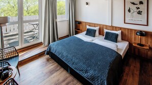 Hotellihuone, jossa on suuri sänky, puulattia, näkymä ulos liukuoven kautta, pieni pöytä tuoleineen ja seinällä oleva kehystetty kuva.