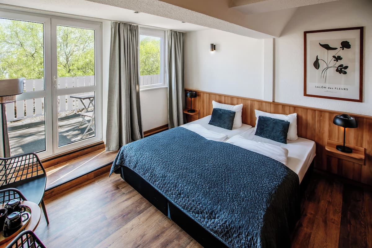 A hotel room with a large bed, wooden floor, a view of the outdoors through a sliding door, a small table with chairs, and a framed picture on the wall.