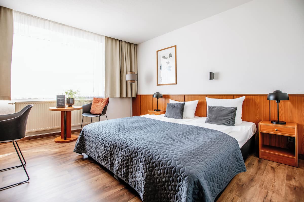 A hotel room with a large bed, a desk, a chair, a nightstand, a lamp, and a window with curtains.