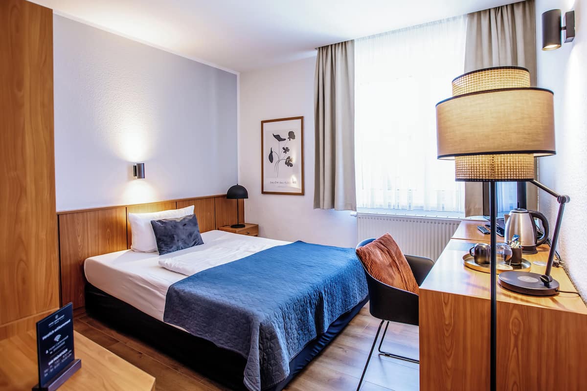 A hotel room with a bed, bedside table, lamp, and a desk with a kettle.