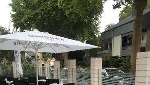 Et udendørs siddeområde med fletstole og borde, parasoller og udsigt til en gade med biler og bygninger.