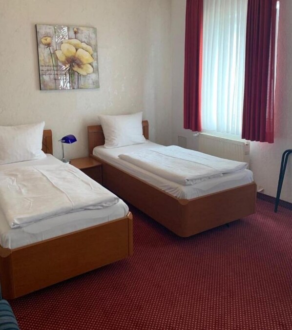 A hotel room with two single beds, a nightstand with a lamp, a painting on the wall, and a window with red curtains. - Hotel Germania (Cologne)