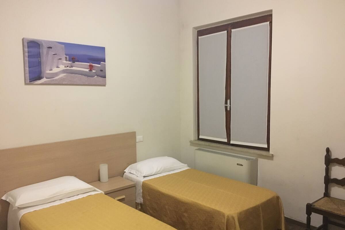 A room with two single beds, a wooden headboard, a window with a view, a radiator, and a painting on the wall.