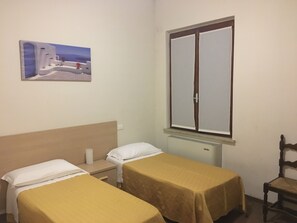 A room with two single beds, a wooden headboard, a window with a view, a radiator, and a painting on the wall. - Blue Hotel Depandance (Suzzara)