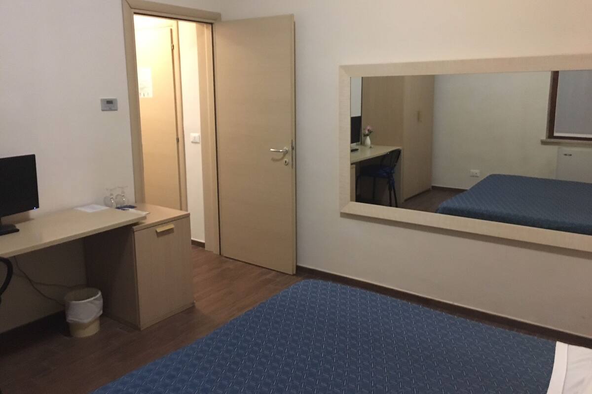 A small, sparsely furnished room with a desk, a chair, a bed, and a door.