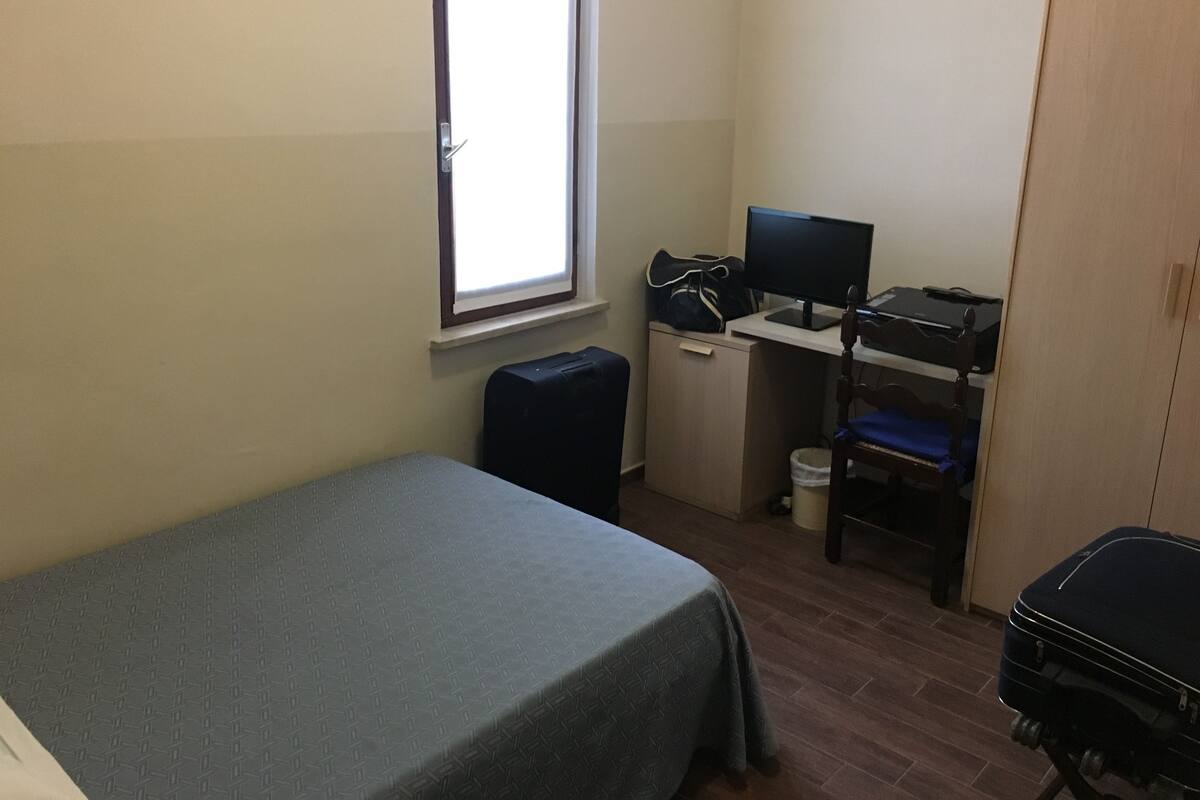 A small, neatly arranged room with a bed, a desk with a computer, a chair, and a suitcase.