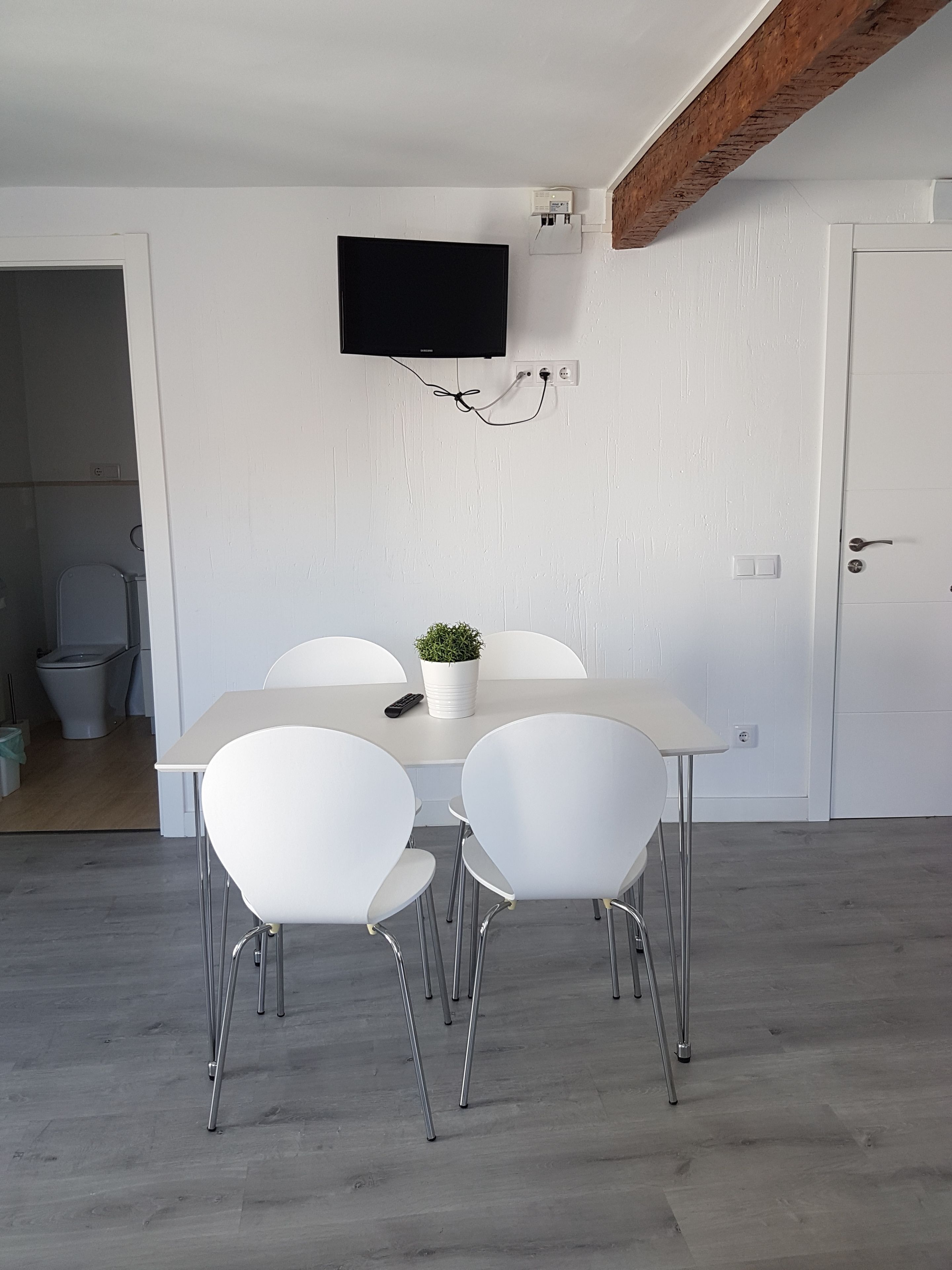 A dining area with a white table and four white chairs, a small potted plant, and a mounted television on the wall.