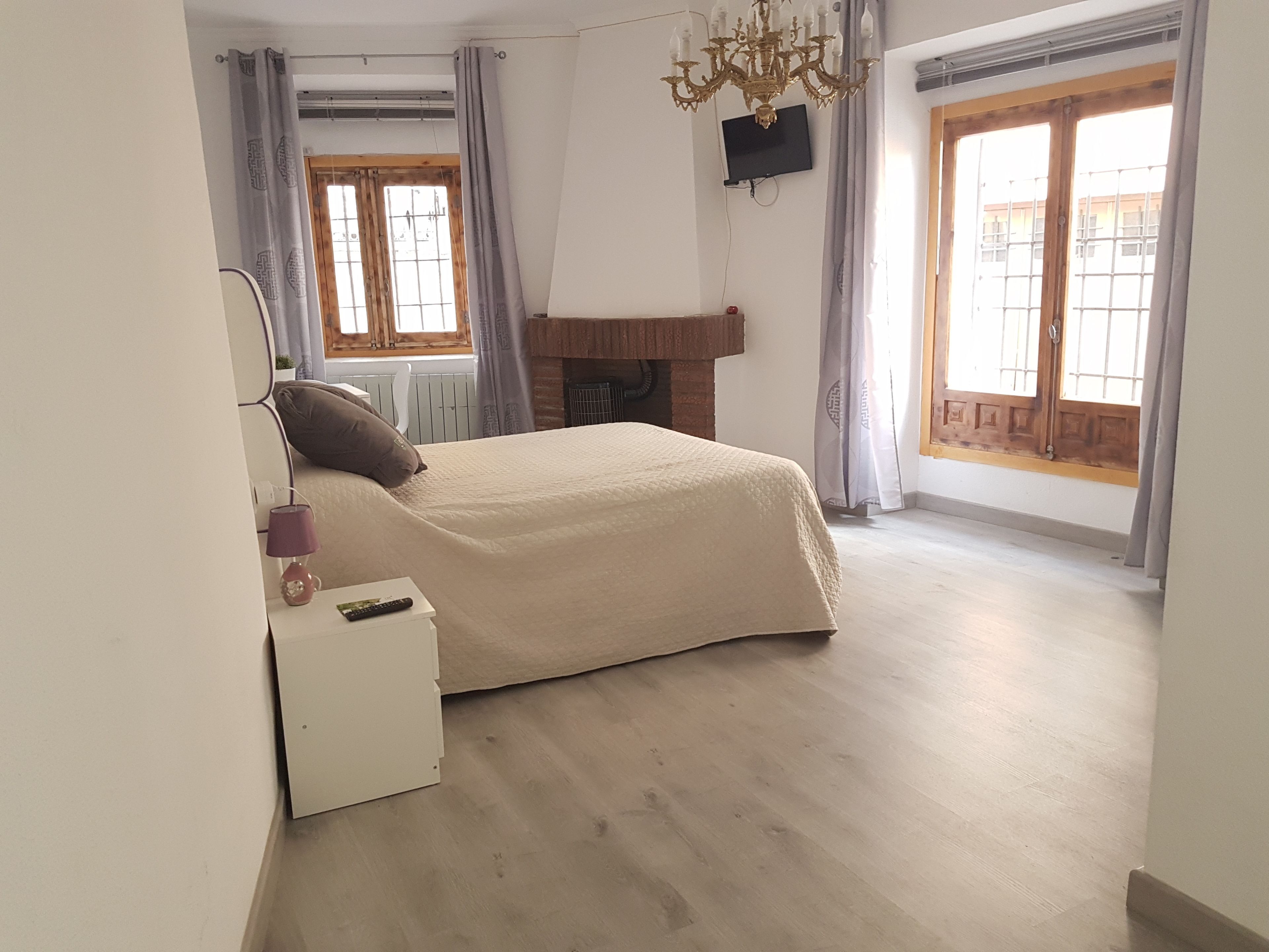 A bedroom with a bed, a TV mounted on the wall, a chandelier, and large windows with curtains.