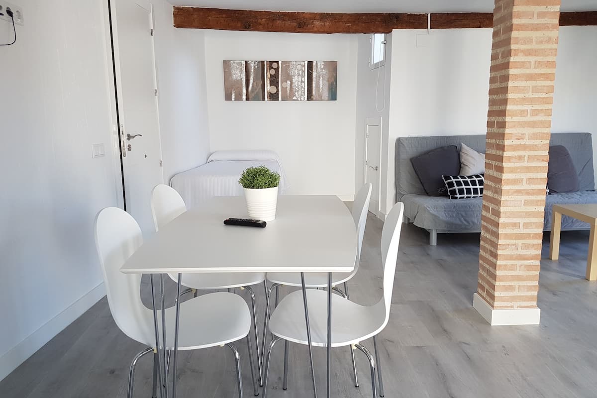 A modern living room with a dining area, featuring a white table, four chairs, a potted plant, and a grey sofa with cushions.