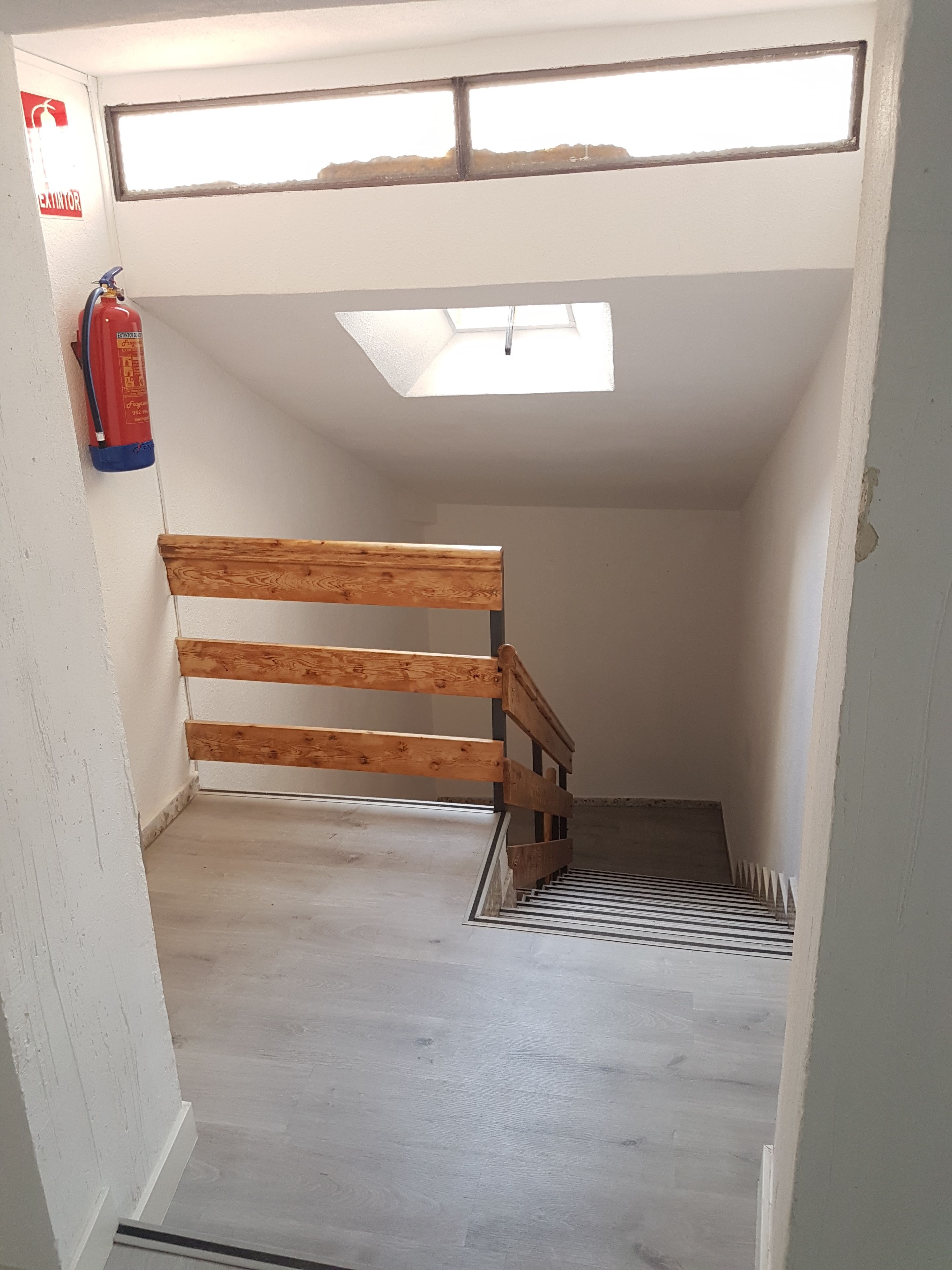 A narrow stairway with a wooden handrail leading to a landing. A fire extinguisher is mounted on the wall to the left.