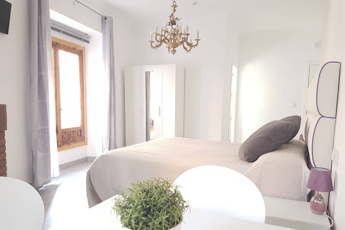A modern bedroom with a large bed, a chandelier, a potted plant, and a view of the outdoors.