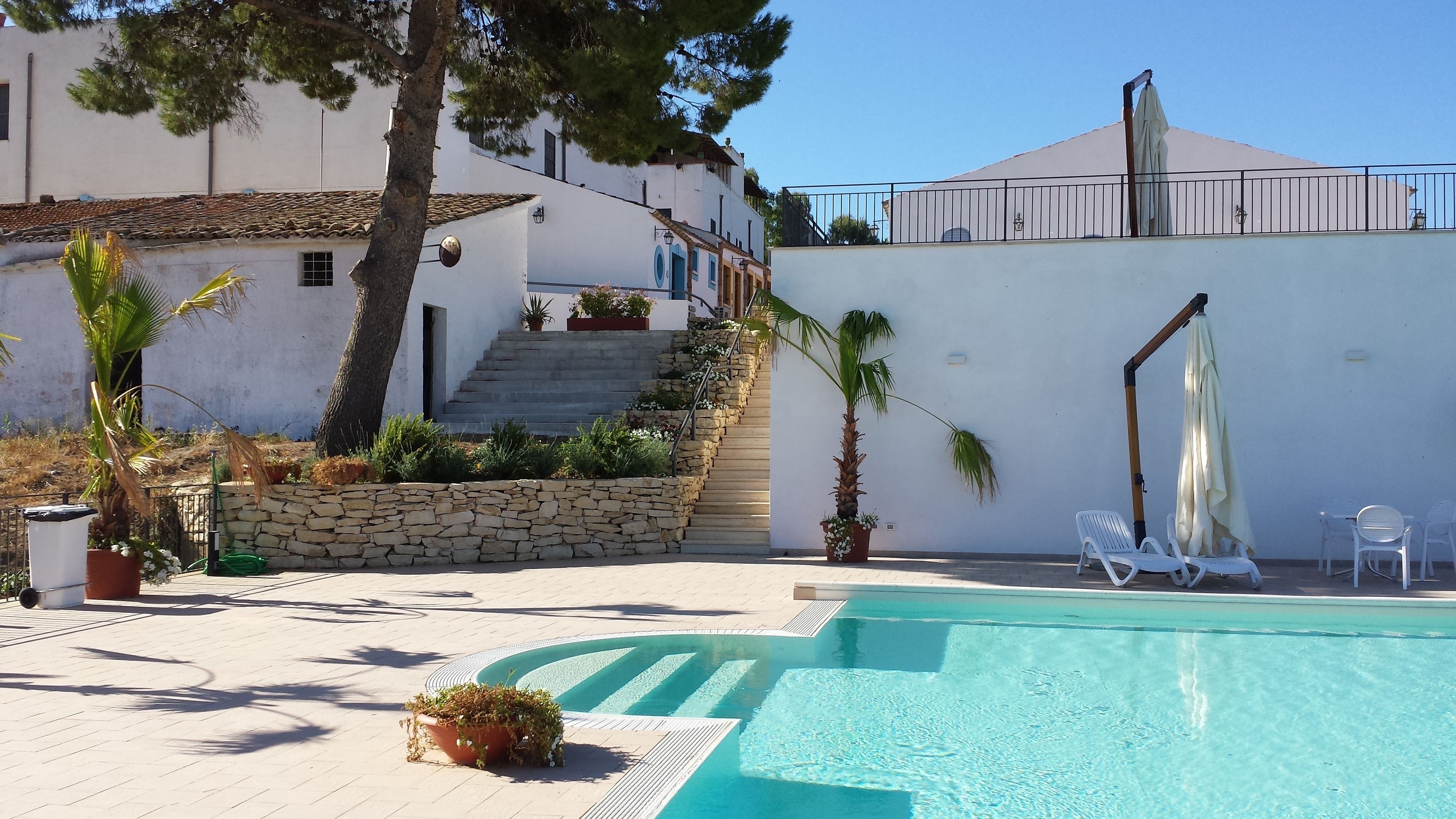 Zona piscina con una scala in pietra che conduce a un edificio bianco, circondata da piante in vaso e un cielo azzurro.