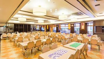 A large banquet hall with rows of tables and chairs, high ceilings with chandeliers, and large windows.