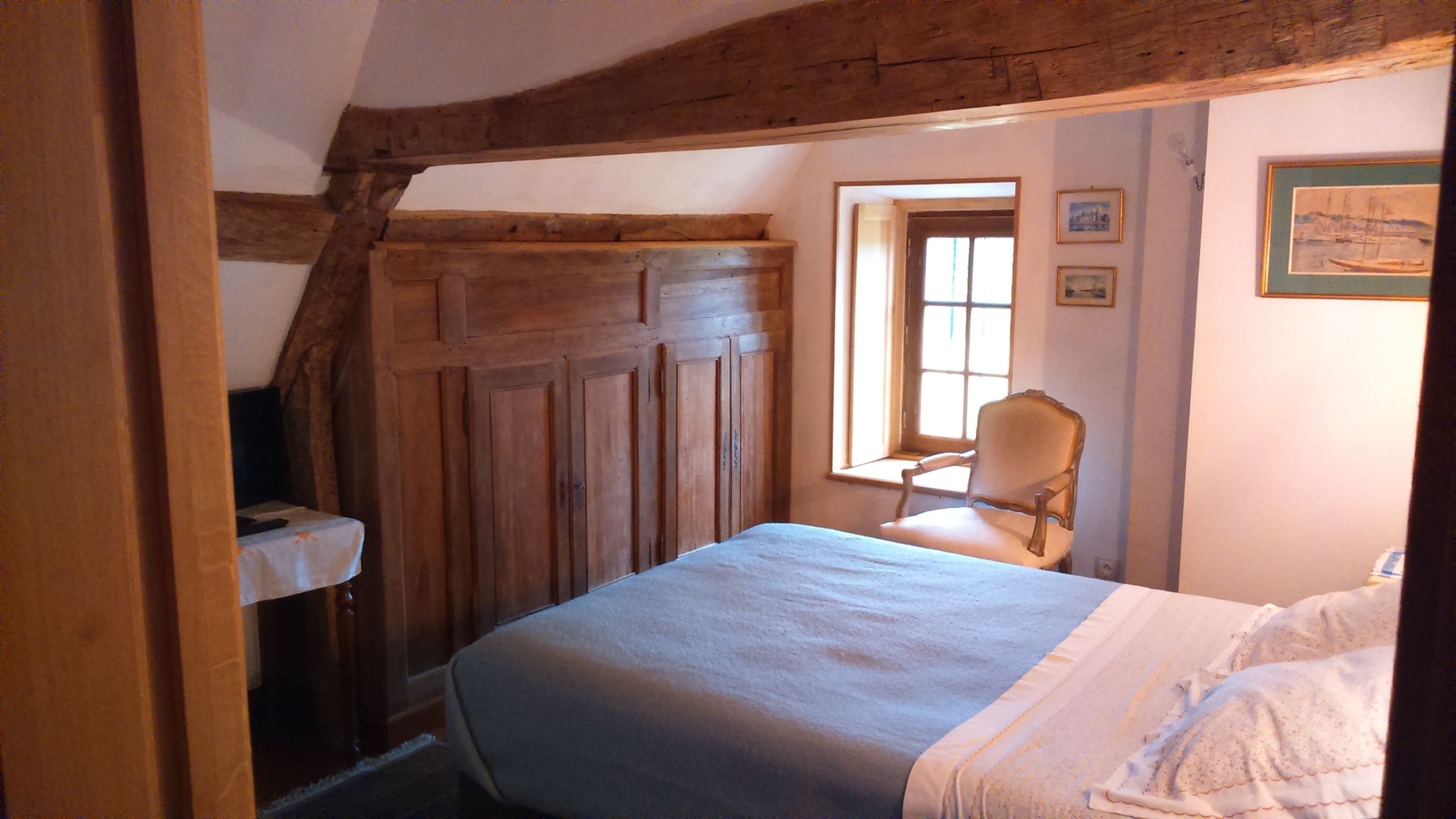 Een slaapkamer met een bed, een stoel, houten balken, een raam en ingekaderde foto's aan de muur.