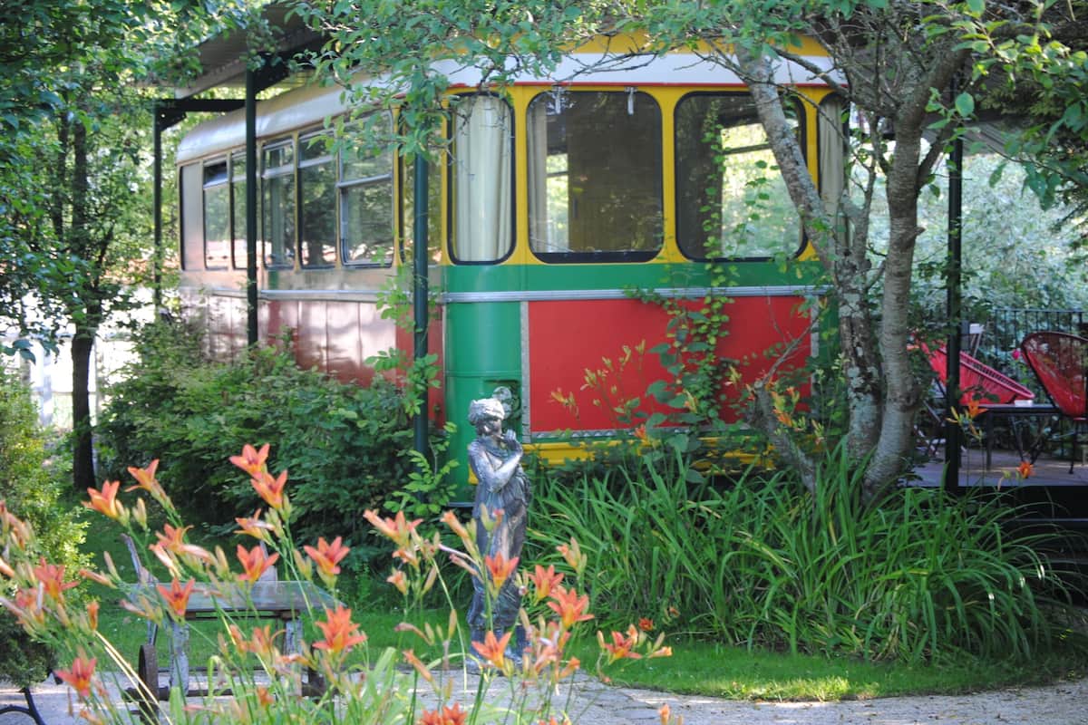 A vintage tram parked in a garden with a statue, surrounded by greenery and flowers.