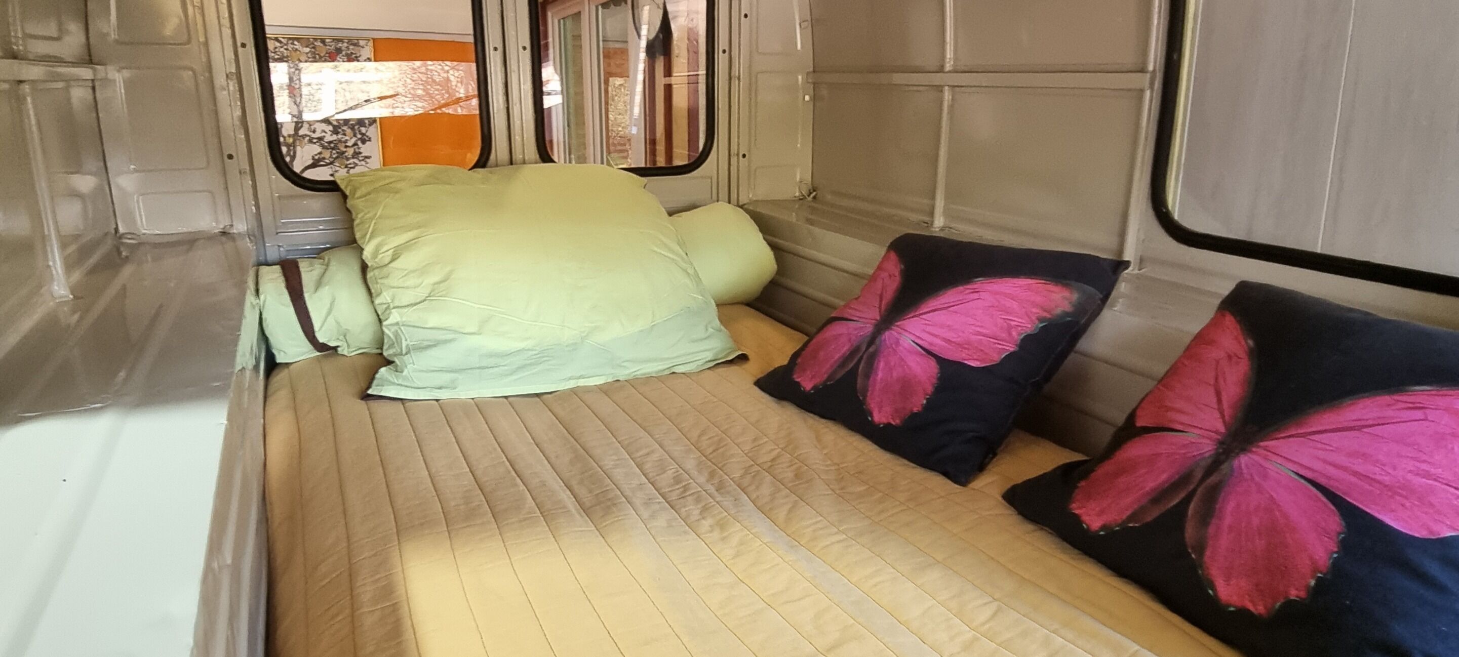 A compact bed with a quilted mattress, two pillows with butterfly designs, and a window with a view of the outside.