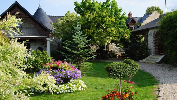 A well-maintained garden with various flower beds and a lawn in front of a house with a stone facade and a pitched roof. - Les Insolites du Grand Logis (Marchais)