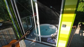 A glass-enclosed hot tub with a champagne bottle on a tray, a guitar, and a wicker chair on a wooden deck. - Les Insolites du Grand Logis (Marchais)