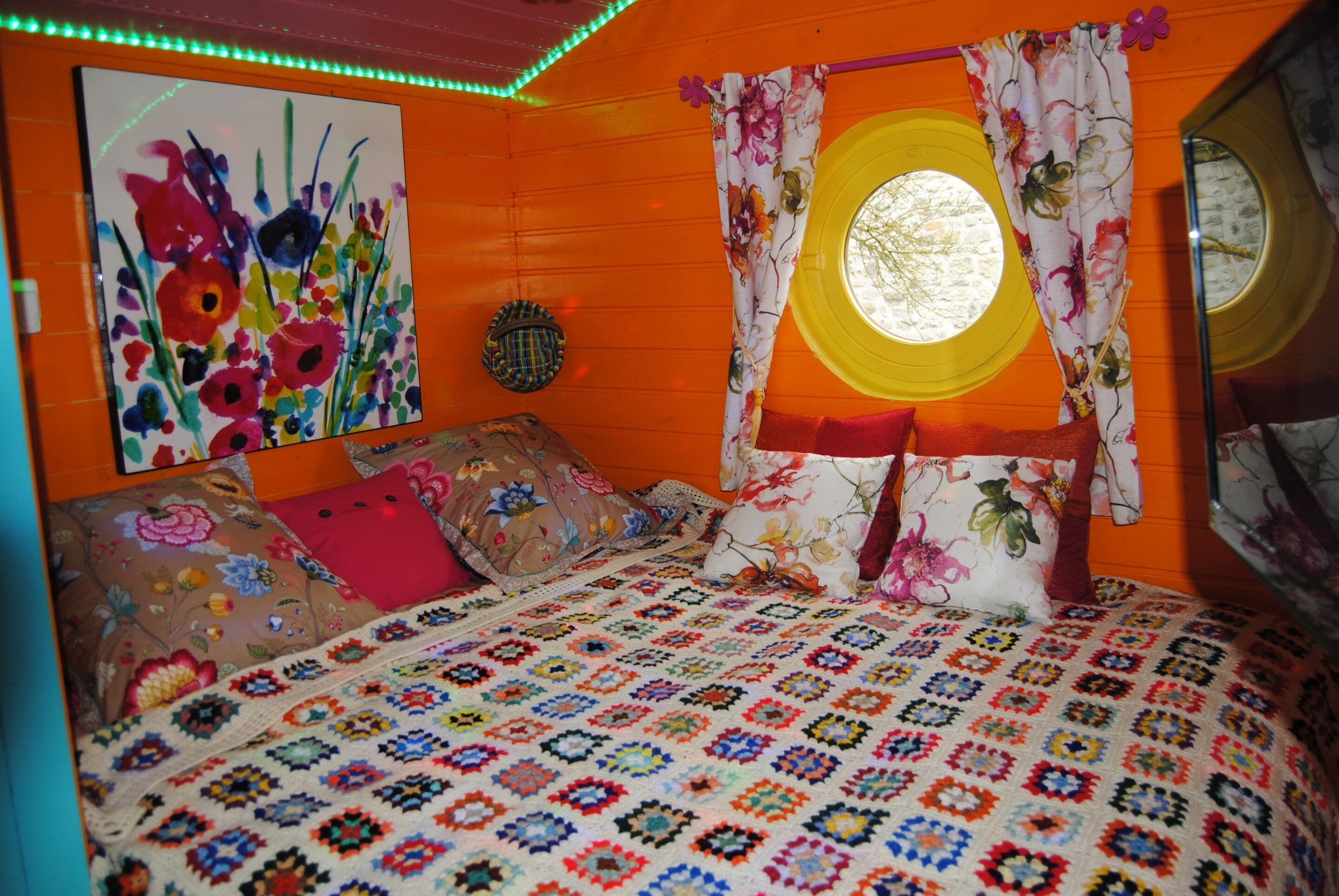 A room with a colorful quilted bedspread, floral curtains, and a round window with a view.
